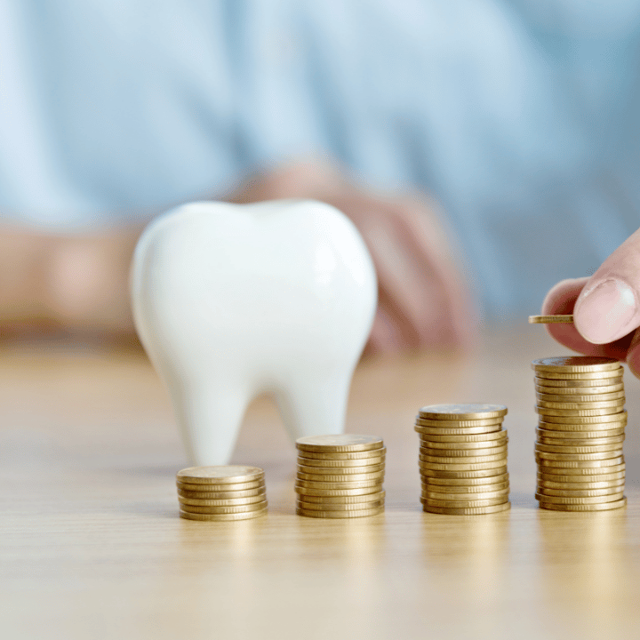 A large white tooth model on a wooden surface next to four stacks of gold coins, with a hand placing a coin on the tallest stack.